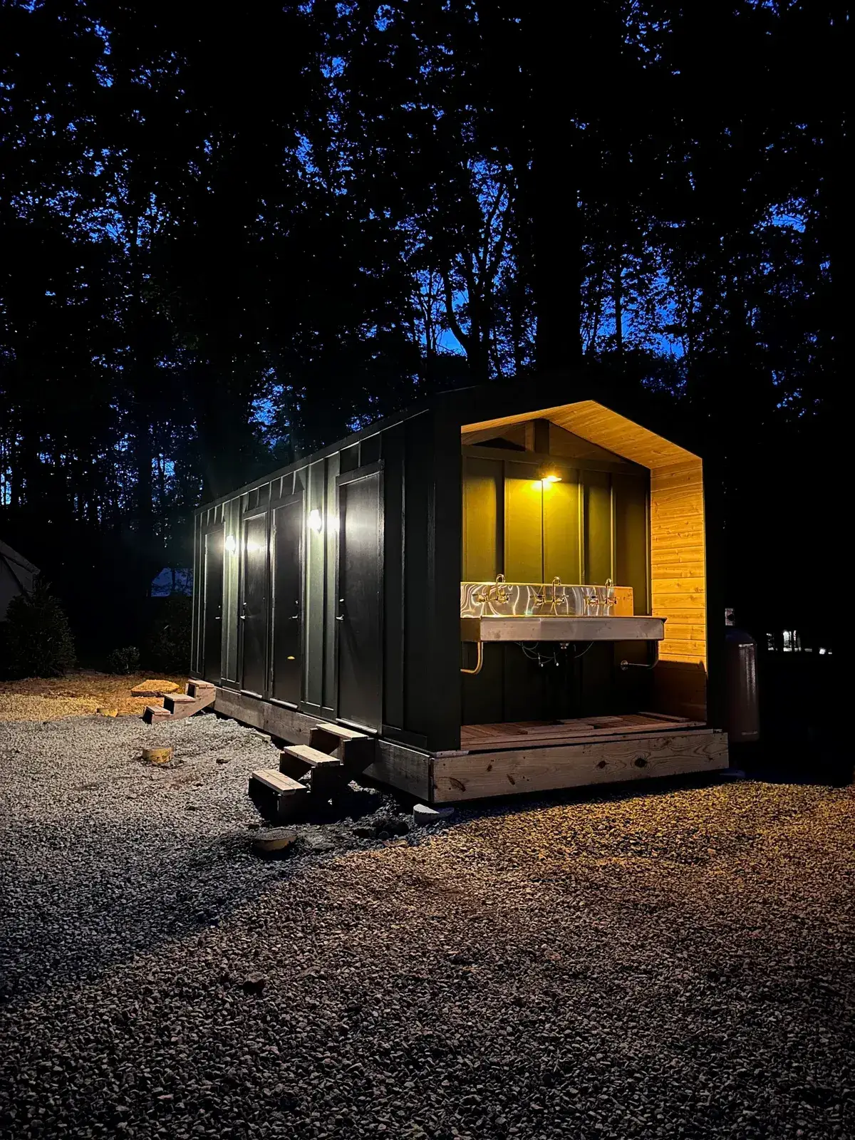 A Pine mobile bathhouse lit at dusk on a wooded gravel pad, delivered as titled, movable equipment